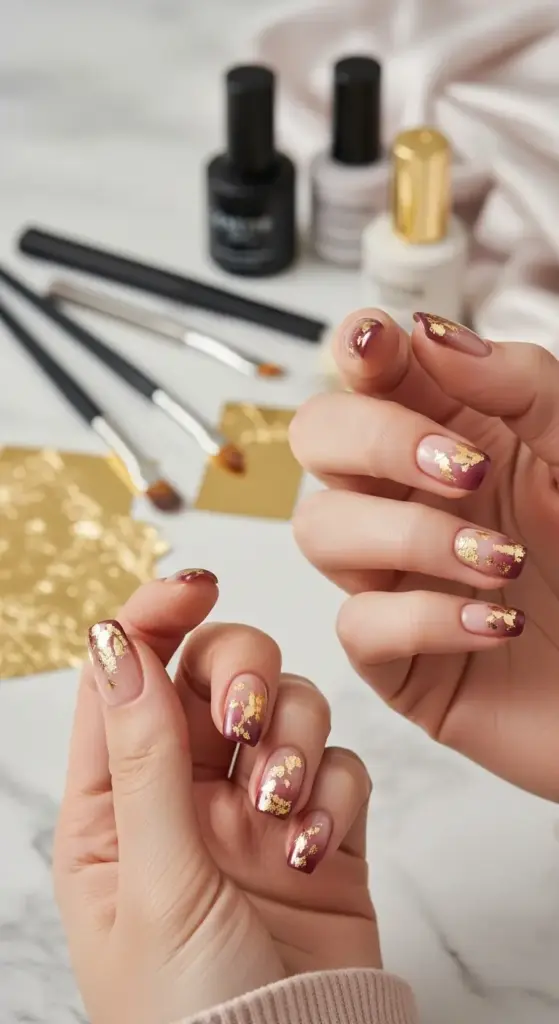Close-up of a stylish manicure with a gold flake design. The nails feature a chic red and gold ombre, with shimmering flakes layered on top. In the background, there are nail polish bottles and brushes on a marble surface.