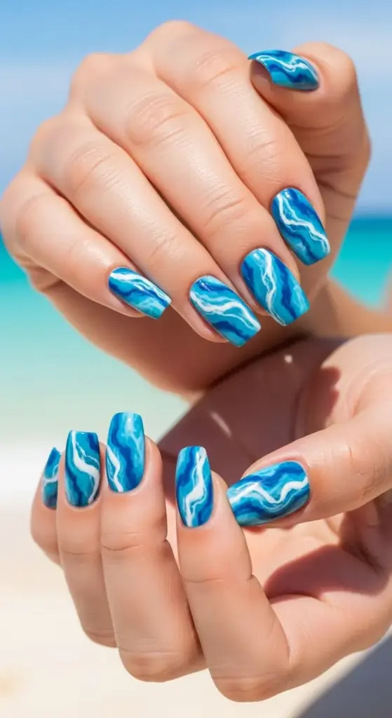 A person's hands are shown against a bright beach background with the turquoise ocean visible. Their nails are painted with a swirling marble design in various shades of blue and white, resembling ocean waves. This eye-catching yet simple beach vacation nail design captures the essence of the sea.