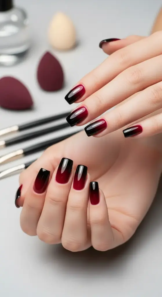 Woman's hands with glossy coffin-shaped dark red nails with a black ombre fade, with nail brushes and makeup sponges in the background.