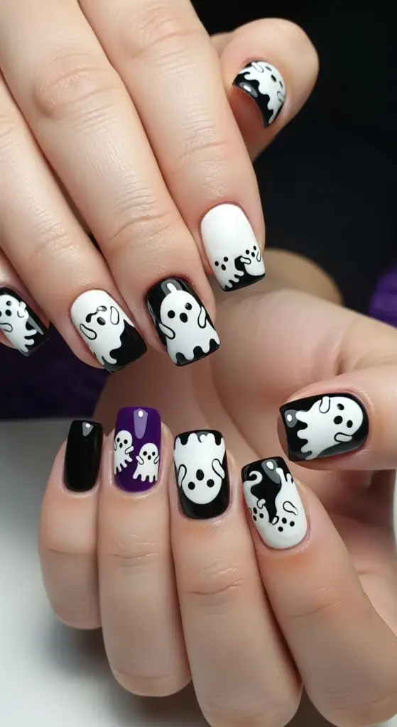A set of cute Halloween nails featuring a playful ghost theme. The nails are a mix of white and black with hand-painted designs. Some nails show black ghosts on a white background, while others show white ghosts on a black background, giving a negative space effect. One nail is a solid black, and another is a vibrant purple with two small white ghosts. The ghosts have a slightly melting or wavy shape, adding a unique, whimsical touch to the manicure.