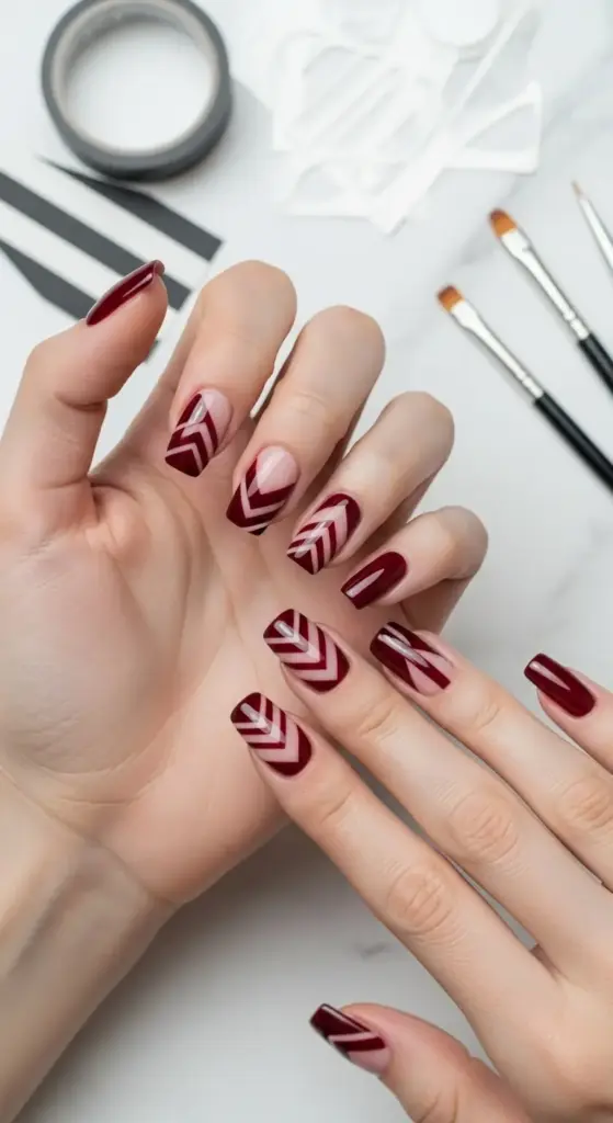 Elegant manicure featuring dark red nails with a chevron and geometric pattern on a few accent nails. The background shows nail art tools and tape on a white surface.