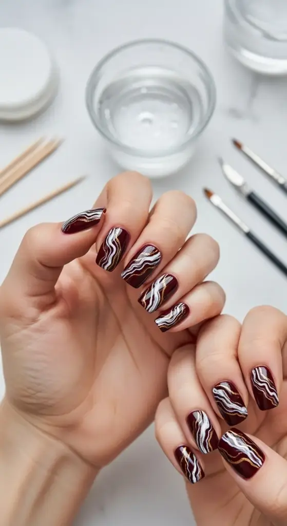 Close-up of a person's hands with glossy, square dark red nails featuring abstract white and gold wavy lines. Manicure tools and a glass of water are visible in the background.